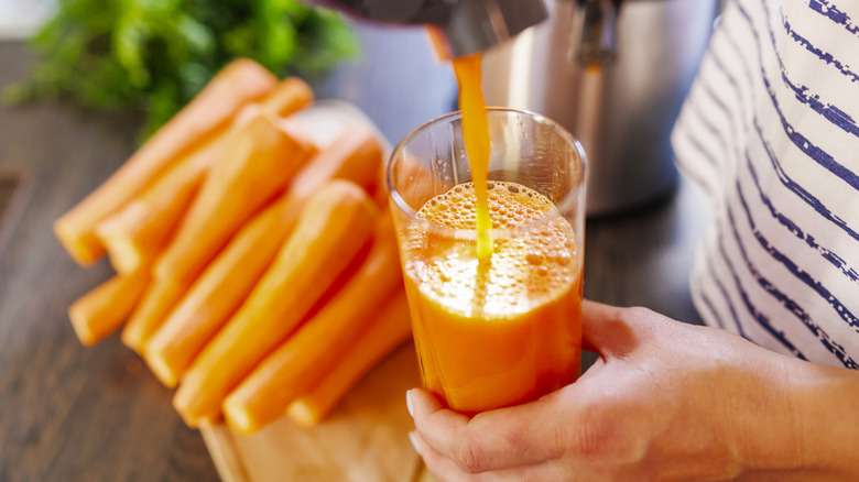 Overhead shot of carrot juice being poured into a glass with blurred carrots on a cutting board in the background.