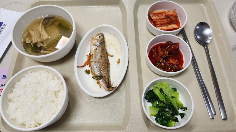 Variety of Korean banchan on hospital tray, including white rice, whole fish, kimchi, and bok choy.