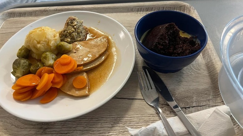 Plate with mixed vegetables and meat, a bowl of cake, and silverware on hospital food tray.