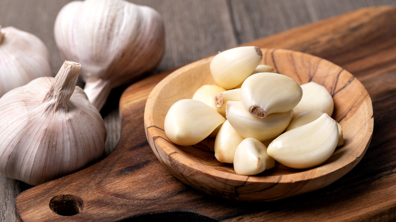 Peeled garlic cloves in a wooden dish on a wooden cutting board with unpeeled garlic heads in the background