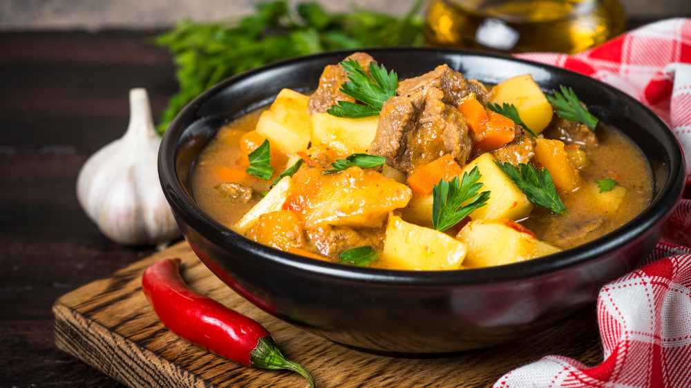 Goulash with meat and vegetables in black bowl on cutting board. Garlic, red pepper, and red-and-white checkered cloth beside bowl.