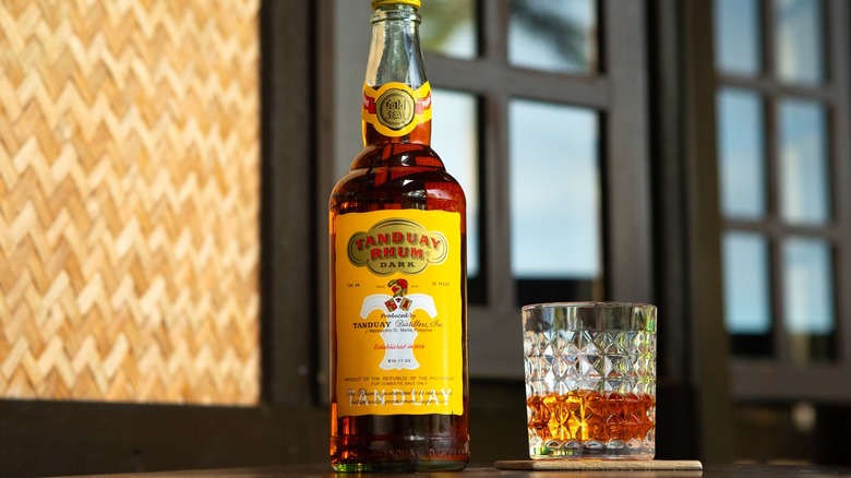 Tanduay Rhum in bottle and glass on wooden table with wall and windows behind them.