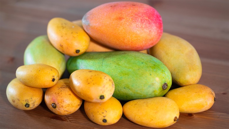 Multiple varieties of mangoes stacked atop each other on a wooden table.