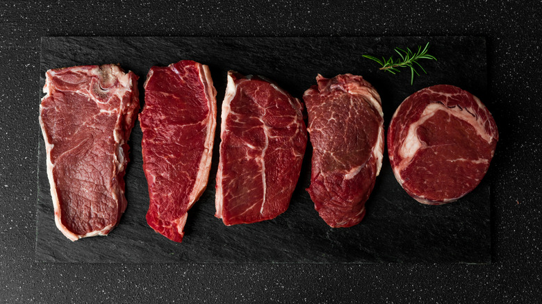 Various cuts of raw steaks on black slate with rosemary sprig.