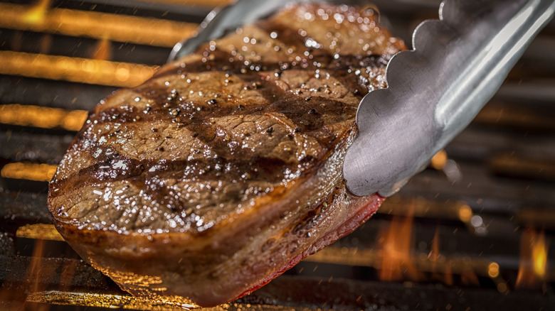 Tongs holding grilled steak over flames.