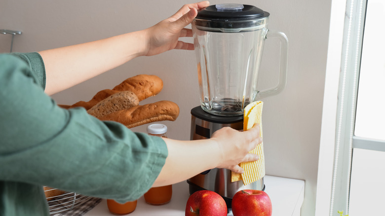 Someone wipes down a blender sitting next to apples and bread on a kitchen counter.