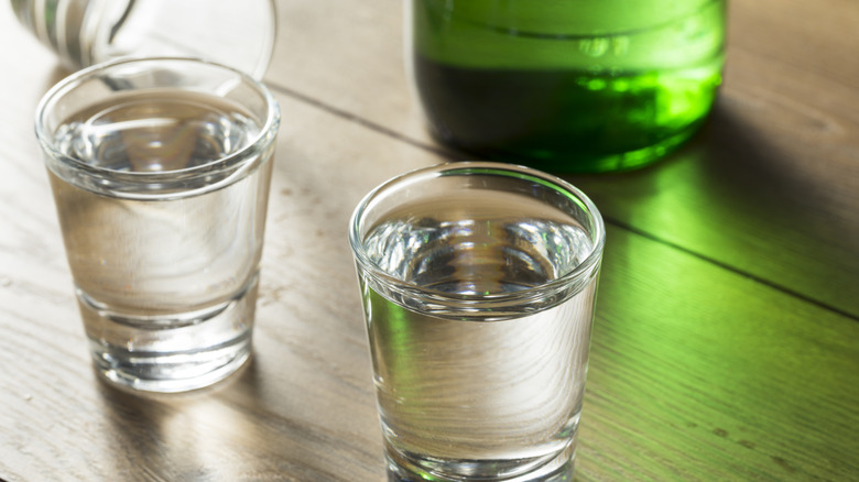 Shot glasses of clear liquid with green bottle in background.