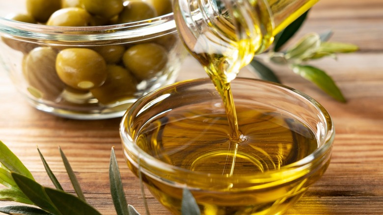 Olive oil in a small, glass bowl with a stream of oil being poured in from above. There are four olives next to the bowl on the left side with olive leaves in the background.