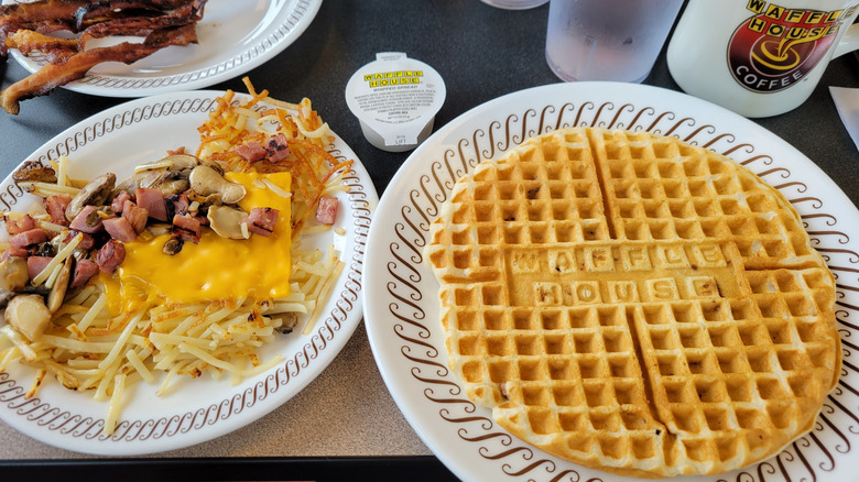 A spread of Waffle house food, including a waffle, hash browns, and bacon plated on the table
