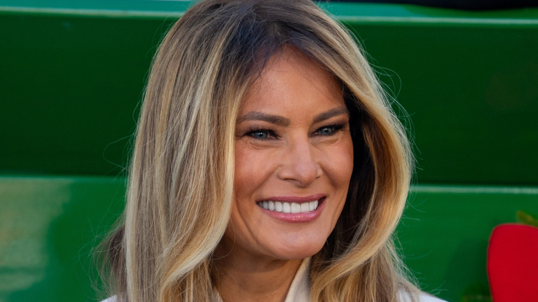 Melania Trump smiling in front of a green oversized box with a red bow
