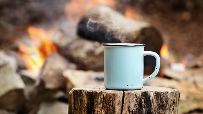 steaming cup of coffee on log of firewood near campfire