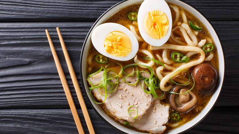 Ramen soup with udon noodles, scallions, egg, mushrooms, pork, served in a bowl next to chopsticks