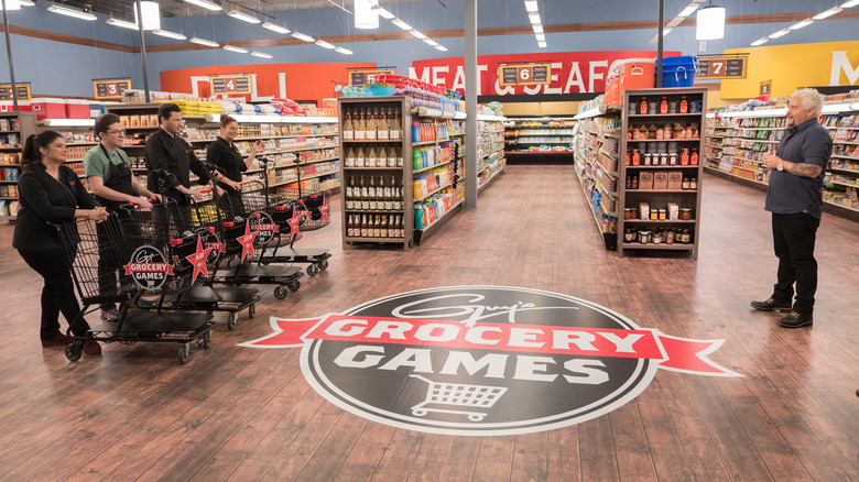 Four contestants stand on the left with carts, Guy Fieri on right standing