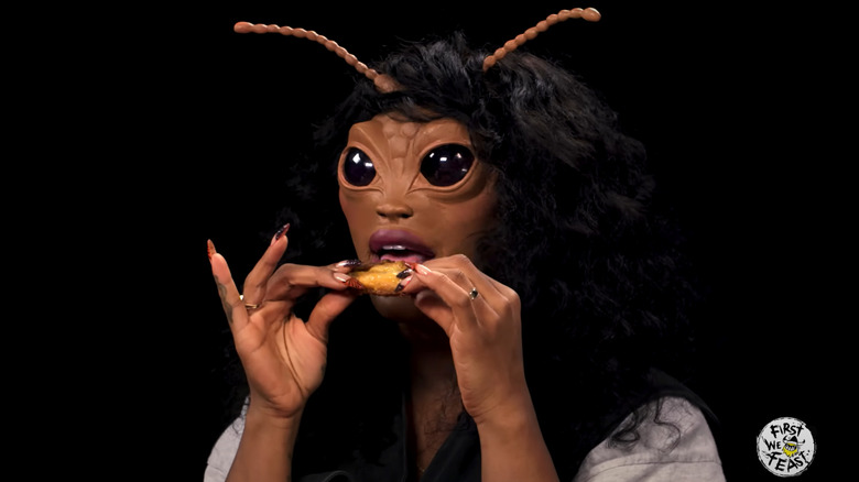 SZA, wearing prosthetics to look like a bug, eating a hot wing