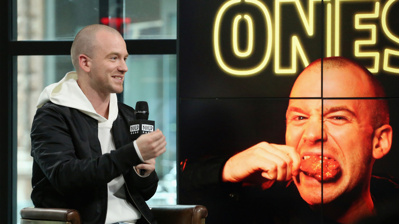 Sean Evans being interviewed, sitting next to a large poster of his own face