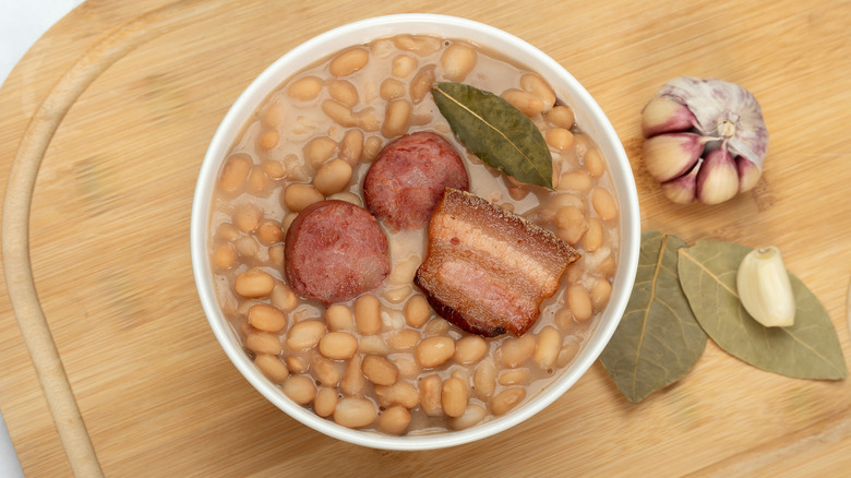 A bowl of pork and beans shot overhead on a wood cutting board next to dried bay leaves and a head of garlic
