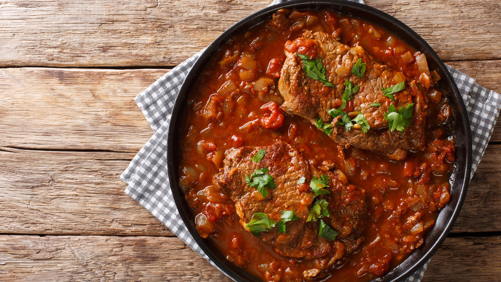 Swiss steak in pan with parsley on top, sitting on grey and white checked cloth on wooden table