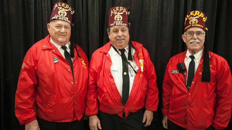 Three Shriner members pose for a photo wearing fezzes and red jackets.