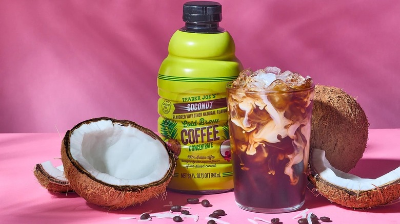 Trader Joe's coconut cold brew concentrate with glass on pink background, coconuts, and coffee beans