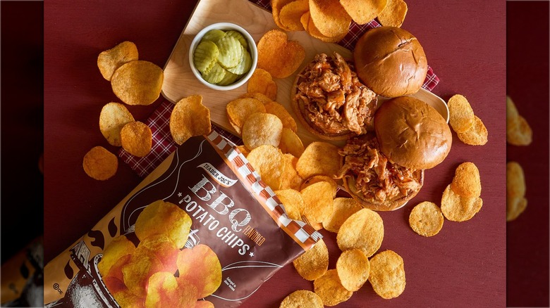 An open bag of barbecue potato chips on table with pulled pork sandwiches and pickles.
