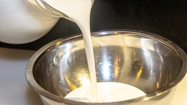 Heavy cream pouring from glass pitcher into metal bowl.