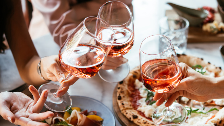 Three people toasting glasses of rosé over pizza.