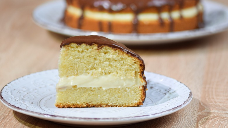 slice of Boston cream pie on a plate with the rest of the dessert in the background