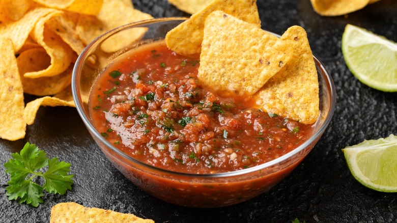 bowl of salsa on a black counter with tortilla chips scattered around