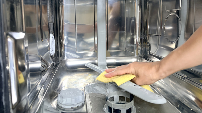 A hand with a yellow cloth cleans the stainless steel interior of a dishwasher