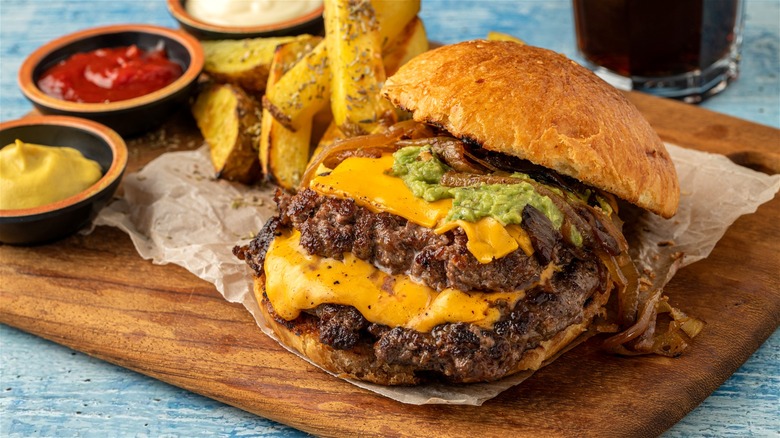 A smash burger with cheese and caramelized onions, served on a wooden board with fries and dips.