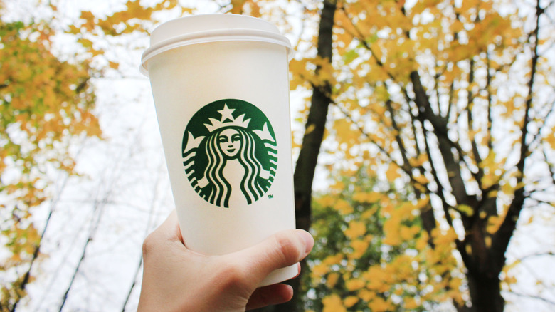 A hand holds a plain, hot Starbucks cup up to trees full of yellow leaves.