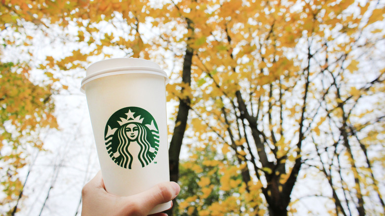 A hand holds a plain hot Starbucks cup up to trees full of yellow leaves.