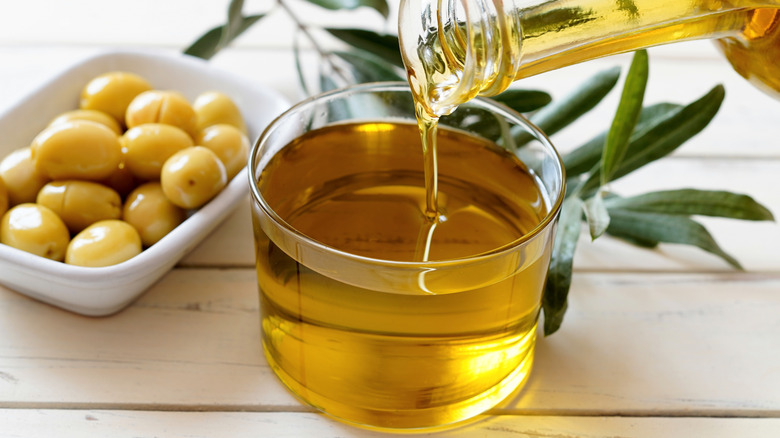 Olive oil being poured into a glass cup with olives in the background.