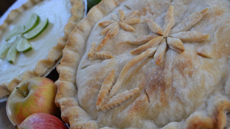 apple pie with flower detail in the crust, next to a key lime pie and apples