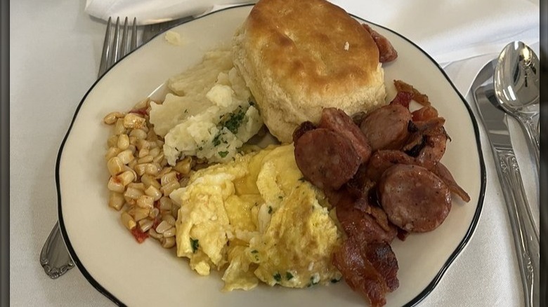 A plate of Cajun sausage and bacon, corn, scrambled eggs, and biscuit.