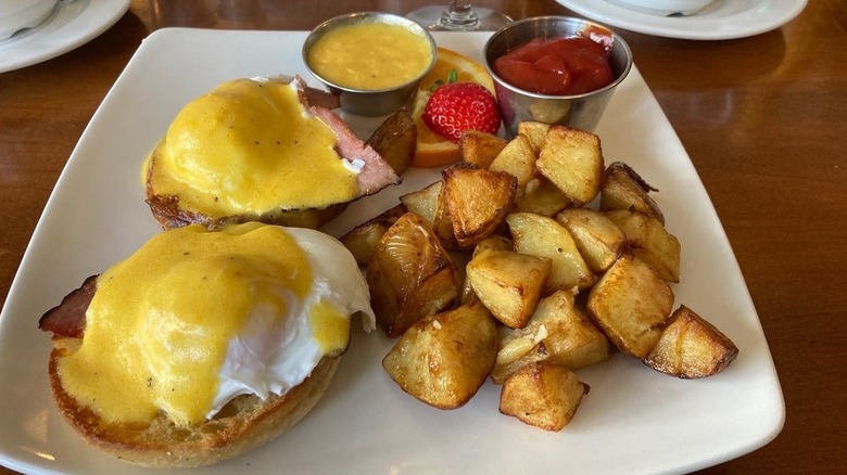 Eggs Benedict, home fries, and fruit on square white plate.