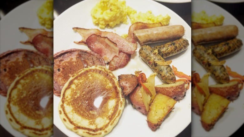 Pancakes, sausage, bacon, eggs, and home fries on white plate.