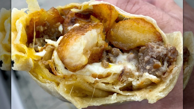 A close-up of a hand holding a breakfast burrito that's been bitten into, stuffed with crispy potatoes, fried eggs, melted cheese, and pieces of maple habanero sausage