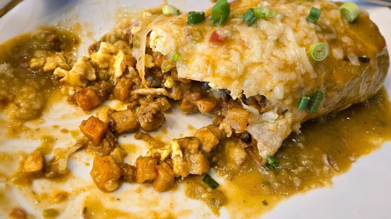 A smothered burrito that's been half-eaten on a white plate with potatoes, eggs, and chorizo oozing out of it