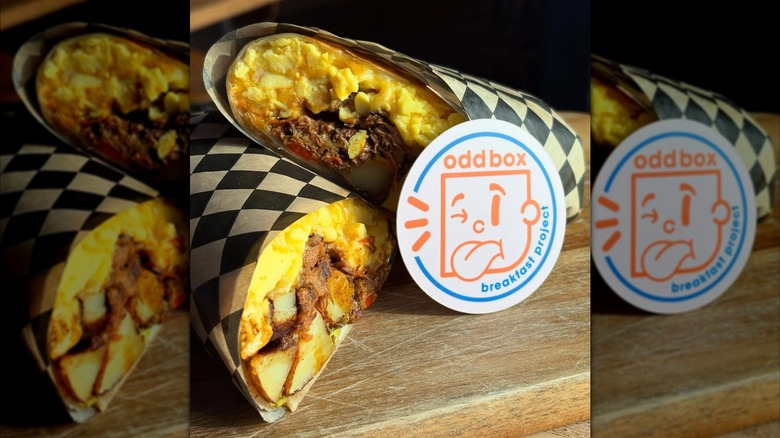 A breakfast burrito wrapped in a black and white checkered food liner and sliced in half. The burrito is stuffed with a potato hash, cheesy eggs, and pork chorizo