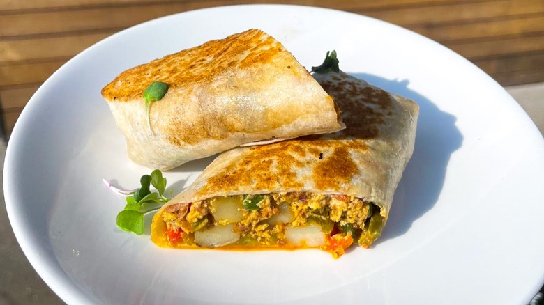 A burrito that's stuffed with scrambled eggs, white cheddar, chorizo, salsa fresca, bell pepper, bacon fat potatoes, and lime crema cut in half on a white plate