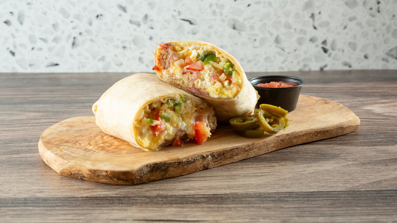 A breakfast burrito filled with eggs, green and red peppers, and ham cut in half on a wooden tray with salsa and jalapeño slices next to it