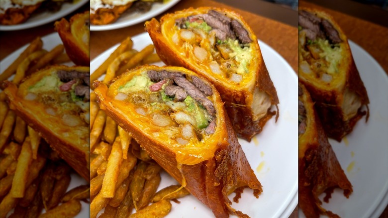 A breakfast burrito cut in half, filled with carne asada, guacamole, sour cream, cheddar cheese, served with a crispy grilled cheese crust and a side of fries
