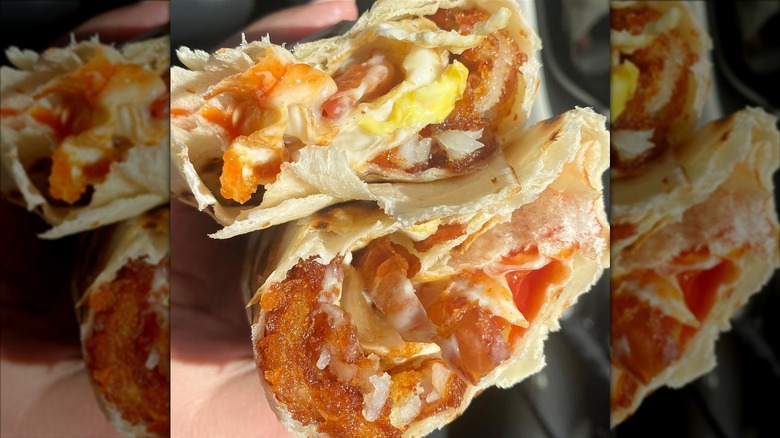 A breakfast burrito cut in half, filled with fried eggs, melted American cheese, bacon, hash browns, and ketchup