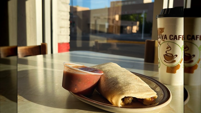 A breakfast burrito on a plate with a side of salsa and a takeout cup of coffee by a window