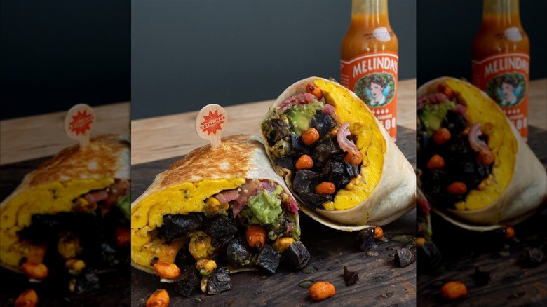 A breakfast burrito cut in half, filled with scrambled eggs, potato hash, avocado, and red onions, served next to a bottle of hot sauce