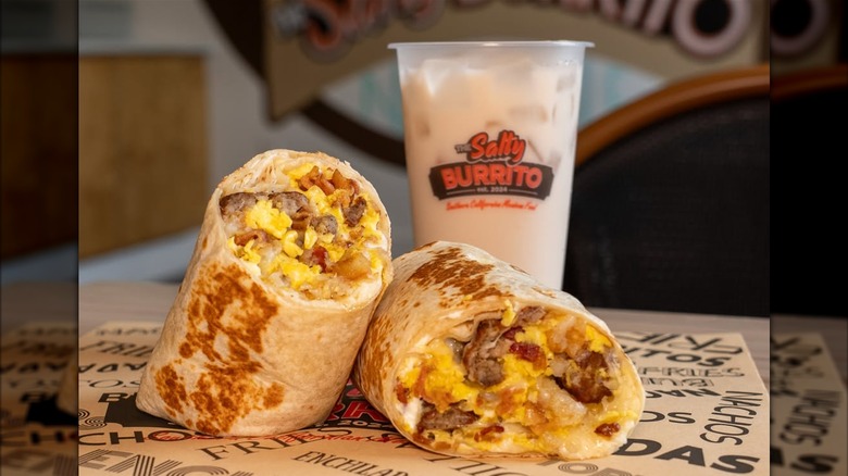 A breakfast burrito cut in half, filled with bacon, sausage, hash browns, sour cream, and cheese, served with an iced drink