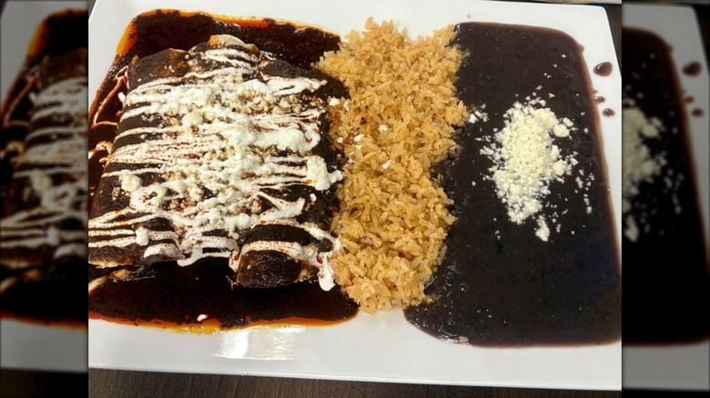 A white rectangular plate with three enchiladas covered in mole, sour cream, and cheese, with rice and beans on the side