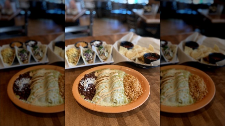 Two enchiladas with green chile sauce with rice and beans on a muted orange plate; tacos and chips and salsa are blurred in the background