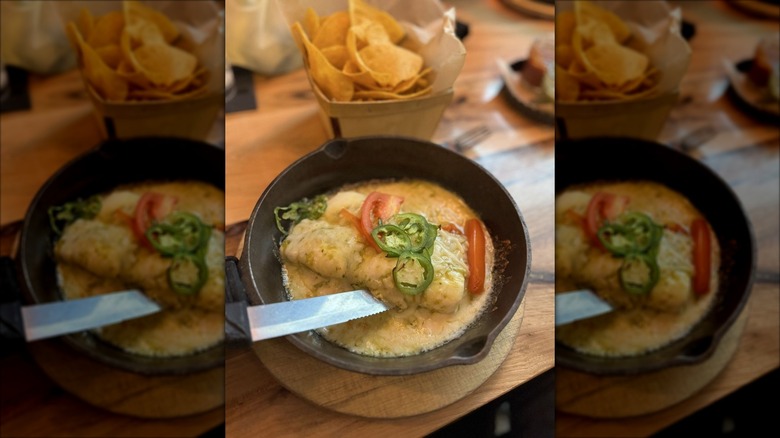 Two cheese-covered enchiladas garnished with sliced jalapeno and tomato in a cast-iron skillet with chips in the background